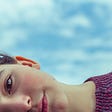 A young woman looks thoughtfully into the camera with a beautiful blue sky filled with fluffy white clouds behind her.