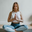 A woman with blonde hair is sitting cross legged in meditation with her hands folded in front of her heart.