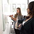 Two women standing at a whiteboard writing on it and working together