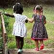 The rear view of two little girls walking hand-in-hand across a timber bridge in a garden. One girl is dark-skinned and wears a white floral dress; the other girl is light-skinned and wears a dark floral dress.