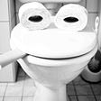 a toilet lid is closed on an empty roll of toilet paper so it looks like it’s smoking a cigar. Two rolls of toilet paper rest on the lid to look like eyes.