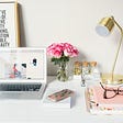 Photo of neat desk with laptop, pink roses, a gold lamp, and pink notebooks.
