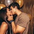 A young, cis het appearing couple kiss in front of some gold and white balloons.