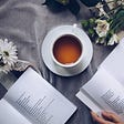 White cup of tea on a grey blanket with daisies and 2 open books nearby