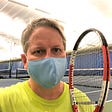 A middle-aged man with blondish hair looks into the camera, taking a selfie while wearing a neon yellow shirt and blue coronavirus mask. He’s holding in his left hand a black and orange tennis racket.