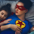 A mother dressed as a superhero is asleep in her bed with a sleeping pre-schooler daughter on one side and a sleeping baby on the other side.