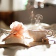 Cup of a hot drink and a pen resting on an open book which is on a bed with sun rays shining on it.