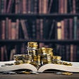 Stacks of coins sitting on top of an open book, on a table with a full bookshelf in the background