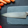 Description: cutting board with knife on top, surrounded by vegetables