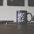 A mug with “hustle” text on it atop a desk.