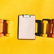 A notepad and with three colorful dumbbells on each side on a yellow background.