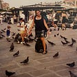 An image of the author in the middle of Piazza San Marco, dressed in shorts, tank top and sandals with my backpack on and my knapsack on the ground, surrounded by pigeons.