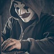An internet scammer working on a keyboard with a mask on his face.