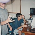 A photograph of Amy smiling at her computer screen, while her brother plays with a balloon.