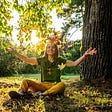 A woman smiles and throws a piles of leaves above her head in the bright sunlight of a park.