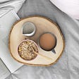 Aesthetic stock image shows a wooden tray with a bowl of oats on it with a pot of milk and a cup of tea. The tray is on a bed with grey sheets and an open magazine next to it. Image is purely for decorative use only to support the blog post on energy levels.