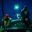 two masked men sit outside a car
