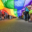 Image description: Many people are lined up holding a very long rainbow banner on a street. The photo is taken from below the banner so only the lower bodies of the people are in the picture.
