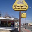 A photograph of the outside of a Winchell’s donut shop. It has a large yellow, triangle shaped sign that says Winchell’s Donut House. There’s a smaller yellow sign that says Open. It depicts a blue sky behind the shop and older model cars on the street.