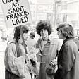 A scruffy young man, dressed in a rough robe, is holding a sign that reads, “Can you live as Saint Francis lived?” This man is holding a pamphet and talking with two young men.