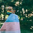 An individual dons a rainbow mask and a trans flag.