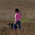 Little girl of color wearing a pink hoody and boots with long pigtails playing in a field with a dog.