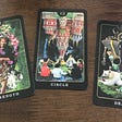 From left to right, three oracle cards — Strength, Circle, and Death are on a dark brown wooden table. Each card shows a collage representing each word. The Strength card shows an elder holding flowers with a tiger. The Circle card in the middle shows people in a circle holding hands with upside-down structures pointing to the people. The Death card shows a dark silhouette with wings standing on top of a white bear that is lying on a large bed of green grass.