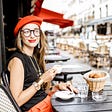 Stereotypical “French girl” in a black top and red skirt with large round glasses, red beret, and lots of artsy gold jewelry sipping espresso in an outdoor cafe with a basket of croissants