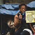 A Black person sits in front of their tent smiling, with their thumb up next to a sign that reads “Mayor Breed we will gladly trade this tent for a hotel room!”