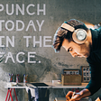 A young man working in an office. On the wall, a sign says: “Punch today in the face” for better productivity.
