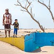 Photo of a father and son holding hands standing on a yellow wall of a house that has lost its roof and has been submerged under sand.