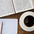 Bible is laid open with a red ribbon bookmarker on a wooden table. Just below the open Bible is a small notebook with a pencil resting on top. Just beside the notebook is a coffee cup and saucer full of black coffee.