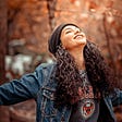 Teenage girl smiling looking up at the sky with eyes closed and arms out wearing a brown beanie and denim jacket.