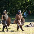 The explorer that stopped 5 minutes before the miracle. nicole wallet. two men dressed in Viking clothing carry shields and swords across a field, reincting a Viking battle.