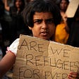 A child refugee holds a sign that says, “Are we refugees forever?”