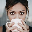 A brown eyed woman looks straight at the camera, with a white mug lifted to her lips, obscuring her mouth.