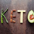 A photograph of asparagus, pecans, cheesem and avocado arranged to spell “keto”.