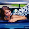 woman with brown hair driving dark blue car polka dot scarf around her neck blowing in the wind