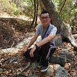 A picture of me in a grey t-shirt and black sweatpants with white stripes on the side crouching in front of a tree at the Rancho Cañada del Oro Open Space Preserve