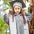 Young preschool girl wearing a grey graduation gown, square academic cap, and stole; holds tassel in hand.