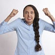 Woman wearing button office style shirt stands in front of a white background with her hands in the air clenched into fists while looking extremely furious