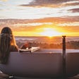 A person with long blond hair sits in an outdoor bathtub, facing away from the camera and toward the sunset on the horizon.