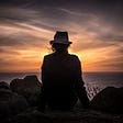 Woman wearing hat sitting on seashore by rocks at sunrise