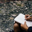 A person sketching a humanoid figure while by a creek. The photo only shows their hands, their sketchbook, and the creek itself.