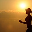 The silhouette of a woman running. In the background, the sun is rising and is covering the sky is a gold hue.