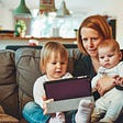 Photo of a woman with two young children, doing a video call on a tablet, sat on a brown sofa.