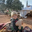 A young child refugee stands in front of a tent, holding a teddy bear.