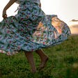 Photo of a woman from the waist down. She is barefoot in a field of grass and her dress is floating as she is dancing and swirling.