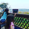Two young girls sitting on a brown deck looking over the railing and holding their iPads on the railing
