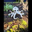 A photo of a small white figurine sitting in a garden patch. The figurine is a skeletal octopus in a fighting stance with tentacles spread wide and glaring skeletal eyes.